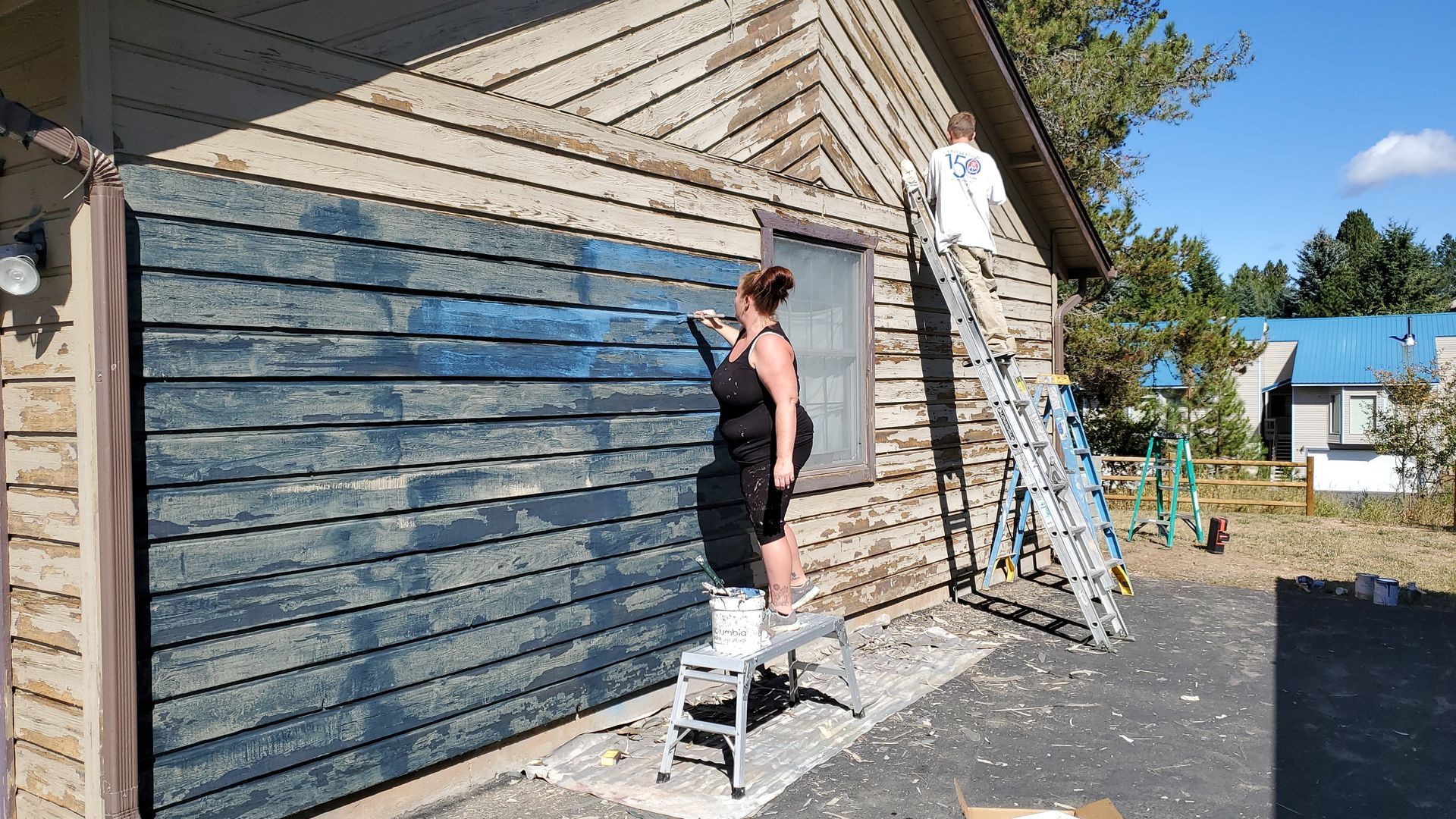 Woman painting horizontal siding blue on a building exterior, standing on a step stool. Another person is on a ladder.