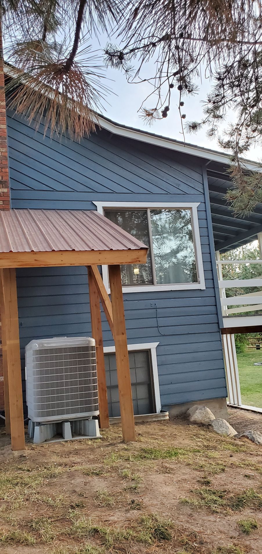 Side of a blue house with a window. A metal roof covers an air conditioning unit and wooden beams. Dry grass surrounds the house.