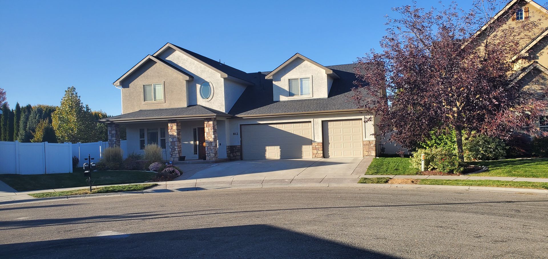 Two-story house with a gray exterior and attached garage, set against a clear blue sky. There is a street and some trees.