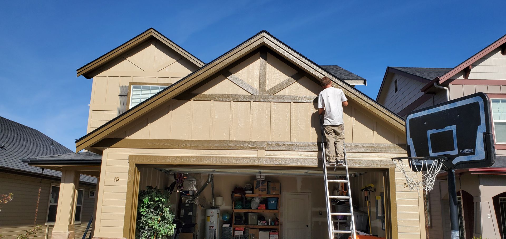 A person on a ladder is working on the exterior of a house. The house has a garage and a basketball hoop.
