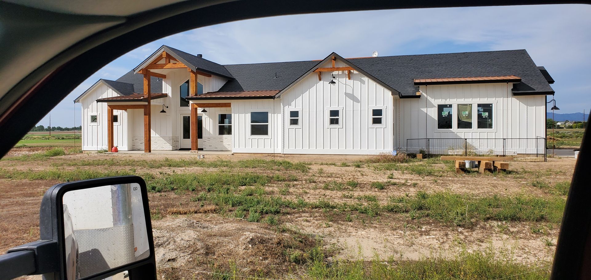 A modern white house under construction, featuring a dark roof and wooden accents. It's set in a field, viewed from inside a vehicle.