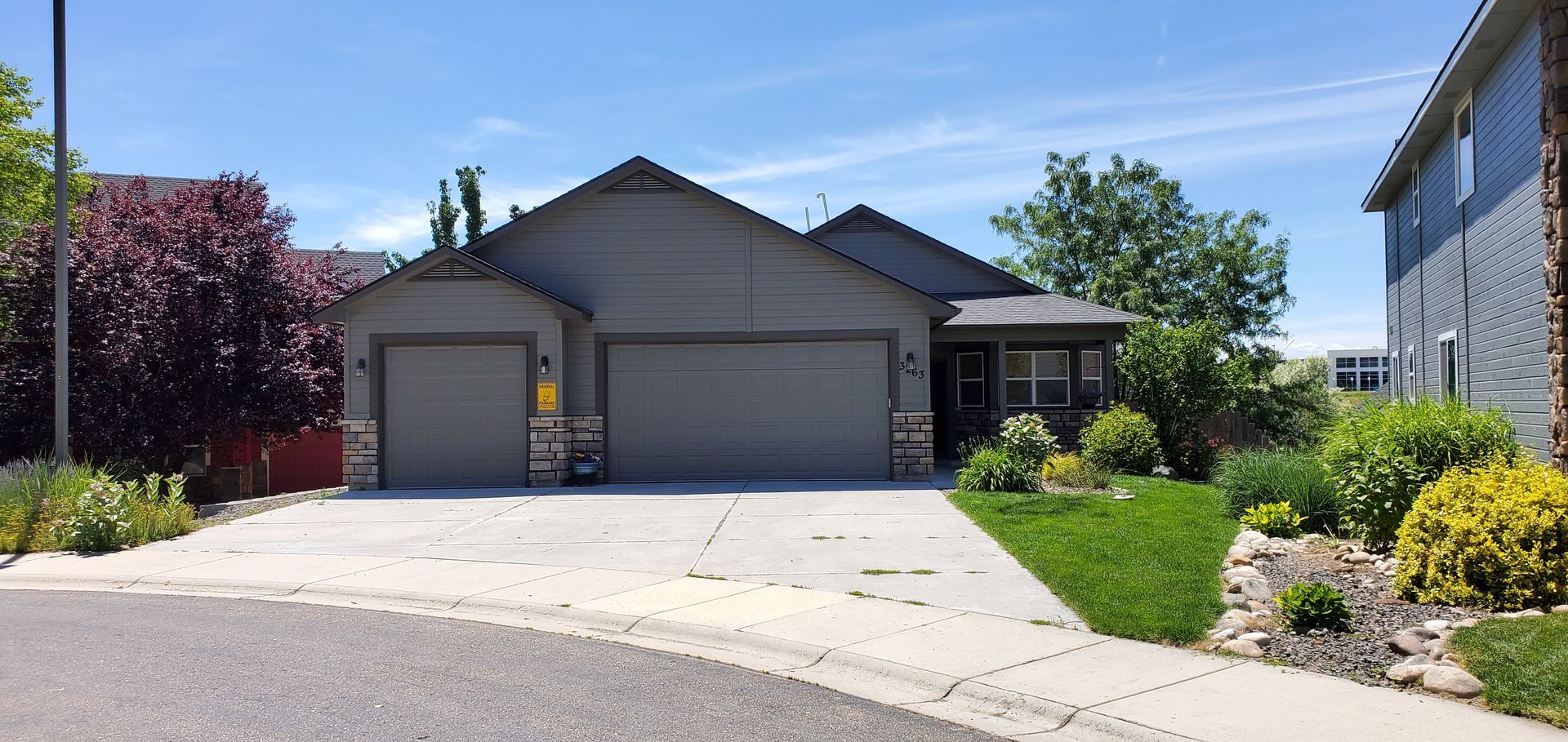 A gray house with a two-car garage sits on a concrete driveway. Green grass and shrubs adorn the front yard. The sky is blue.