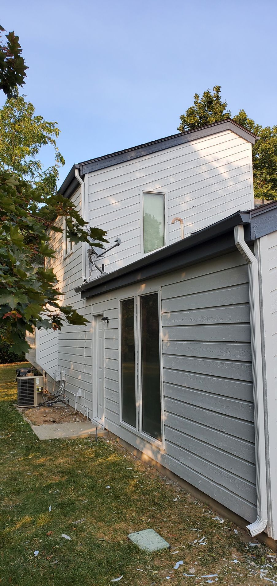 A gray and white house with a small window and large glass doors, surrounded by green grass and trees, on a clear day.