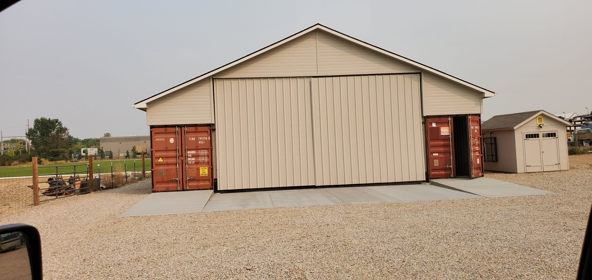 A large building with a tan roof and beige walls. Two opened red shipping containers are on either side of a large door. A smaller white shed is next to the building.
