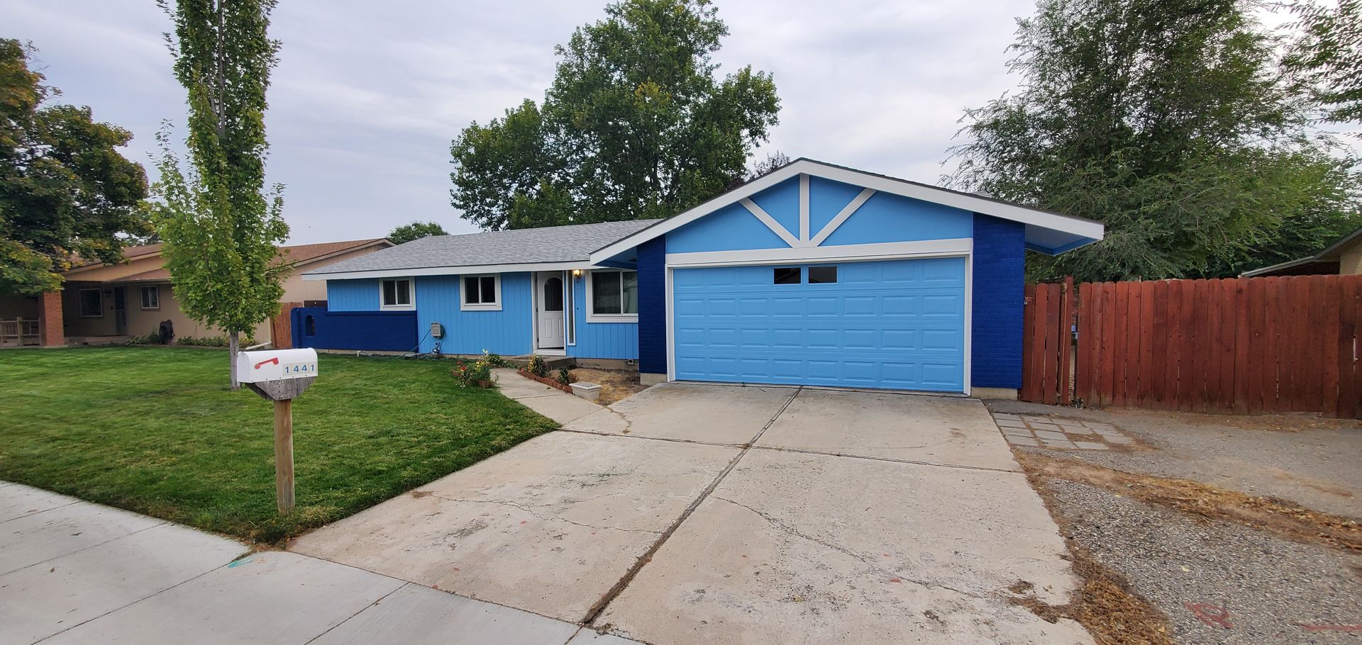 A blue house with a light blue garage door and white trim. A concrete driveway leads up to the house, and a lawn is in the foreground.