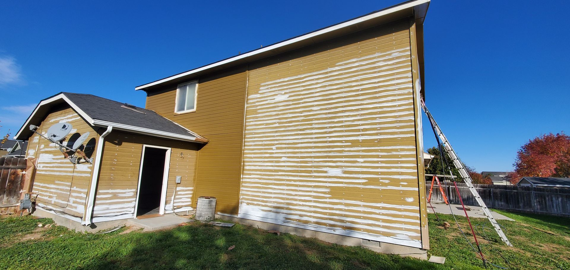 A two-story house with weathered siding and a detached structure; a ladder leans against the side. Blue sky.