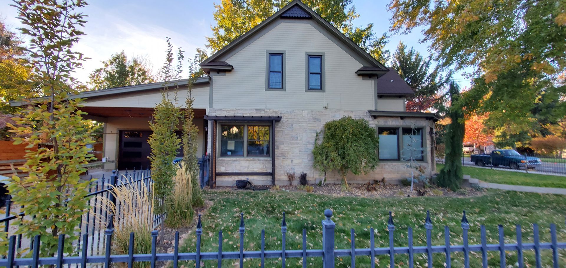 A two-story house with beige walls and dark trim. A black wrought iron fence surrounds the front yard. Green and yellow trees are in the background.