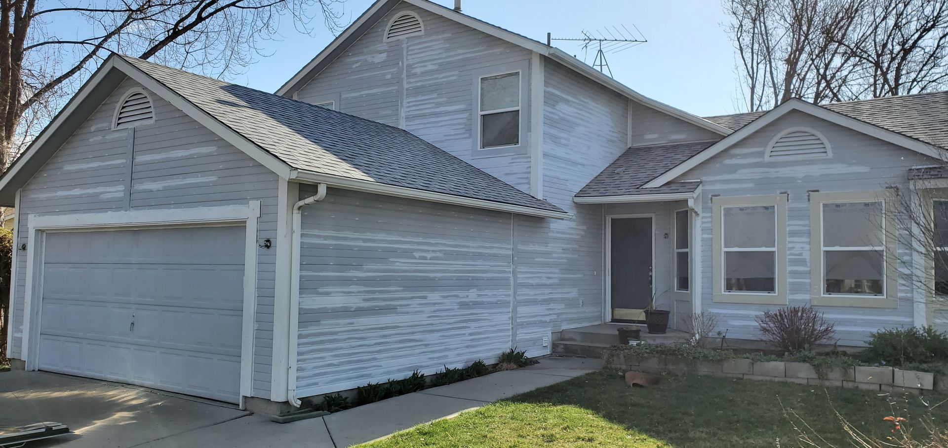 A two-story house with a light blue exterior, gray roof, and a two-car garage door. The house has some visible weather damage.