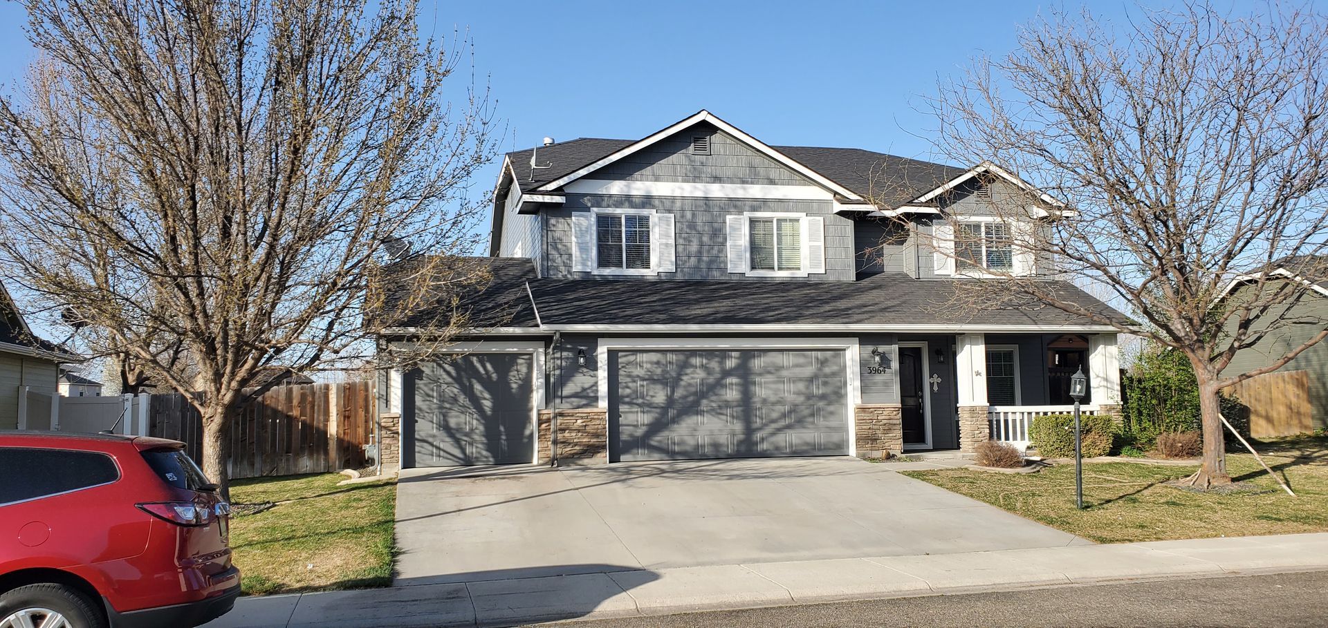Two-story gray house with black roof, a red car parked on the left, and trees with bare branches. Blue sky in the background.