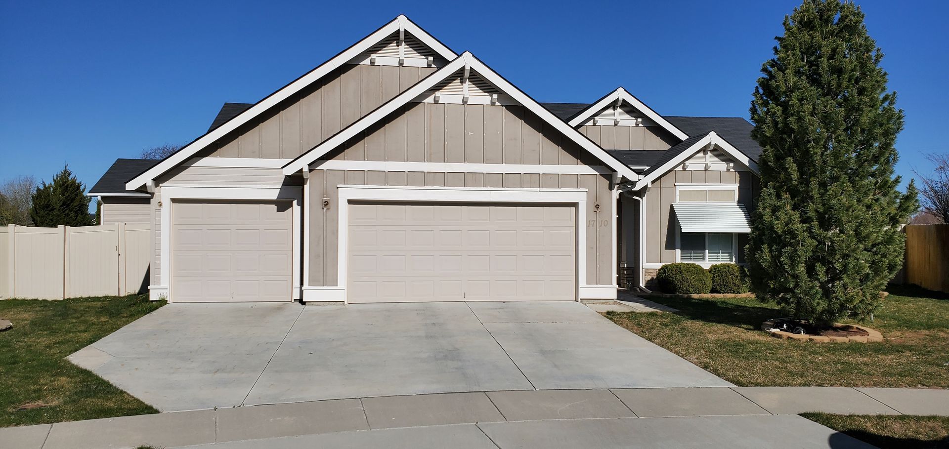 A beige house with a two-car garage and a tall evergreen tree in front, under a clear blue sky.