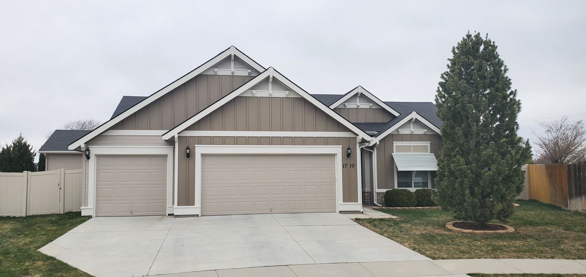 A house with a gray exterior, three garage doors, and a tall green tree in the front yard. The sky is overcast.
