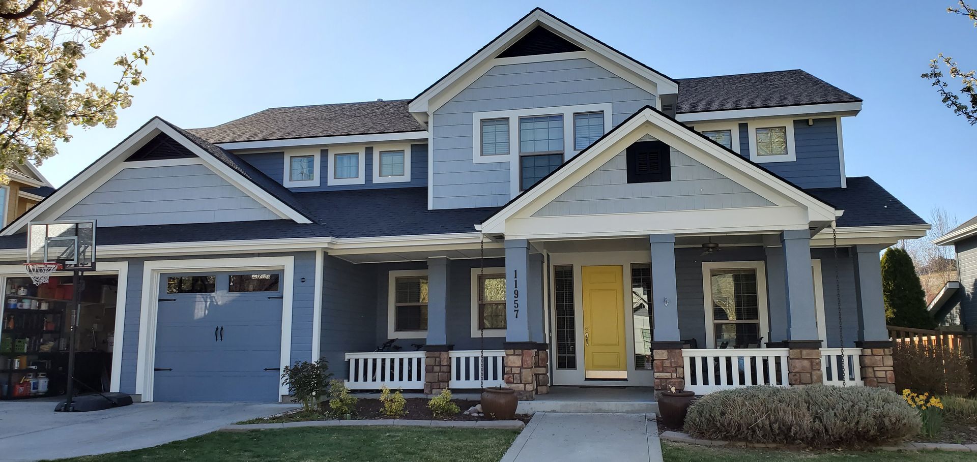 A two-story Craftsman-style house with blue siding and a yellow front door. The house has a front porch, a dark gray roof, and a driveway.