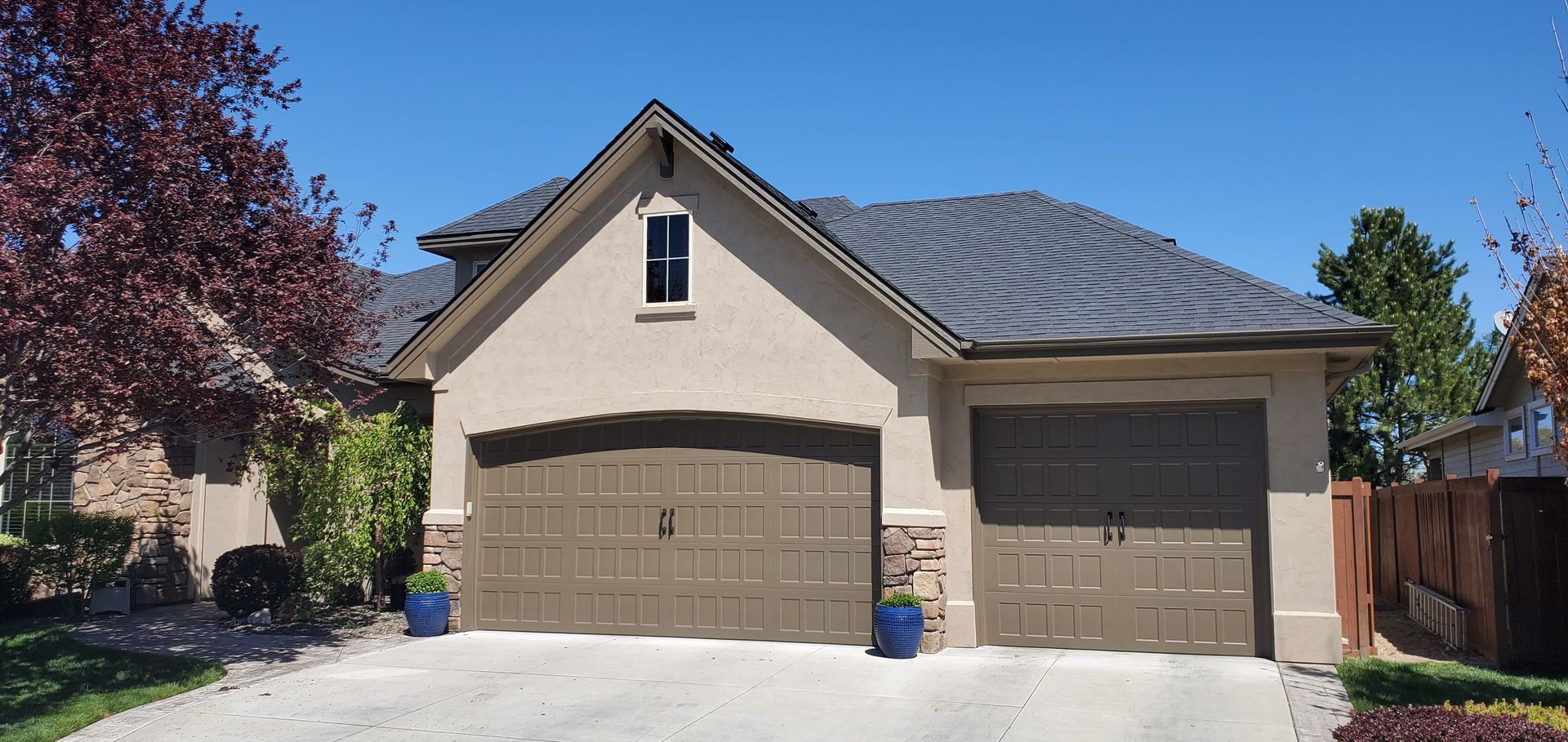 A beige house with a brown garage door on a bright, sunny day. There are bushes and trees near the house.