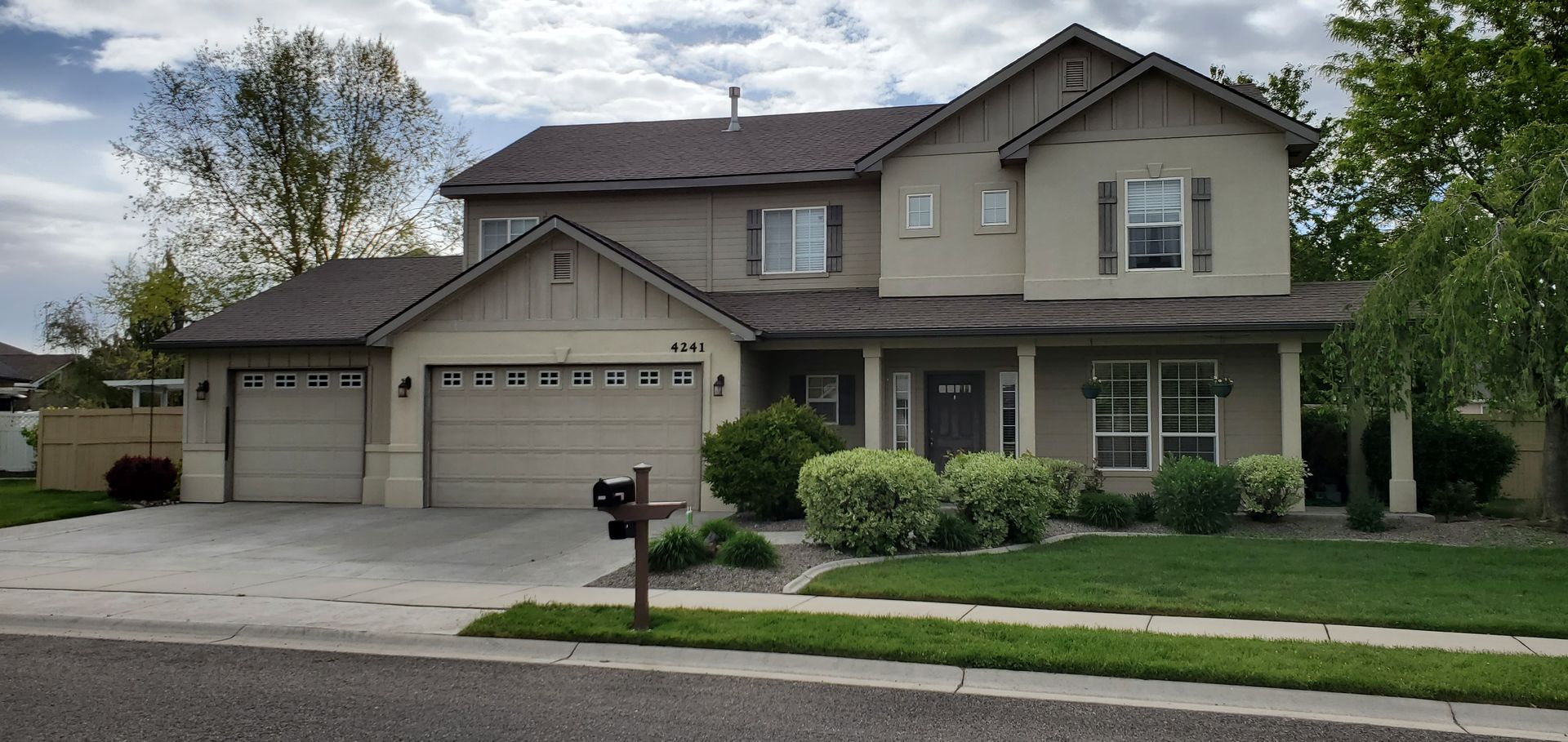 A two-story beige house with a gray roof, a porch, and a three-car garage. The house has a well-manicured lawn.