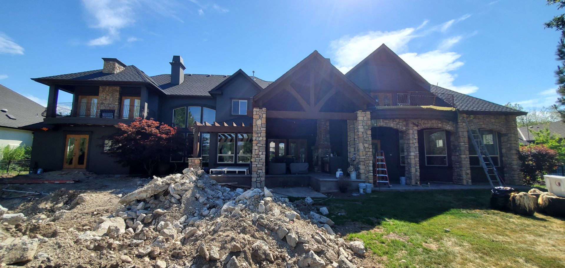 Rear exterior of a large house with a dark facade, stone columns, and a covered patio. Construction debris is piled in the foreground.