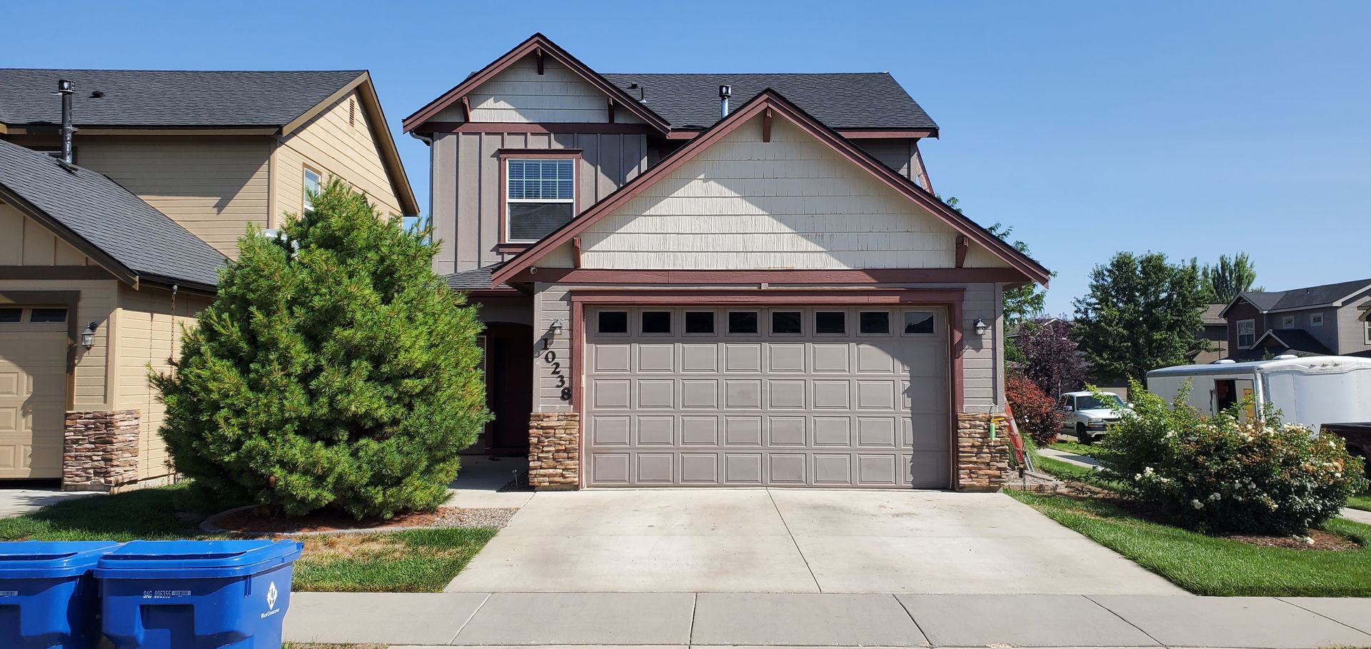 A two-story house with a gray garage door, brown trim, and a small front yard under a bright blue sky.