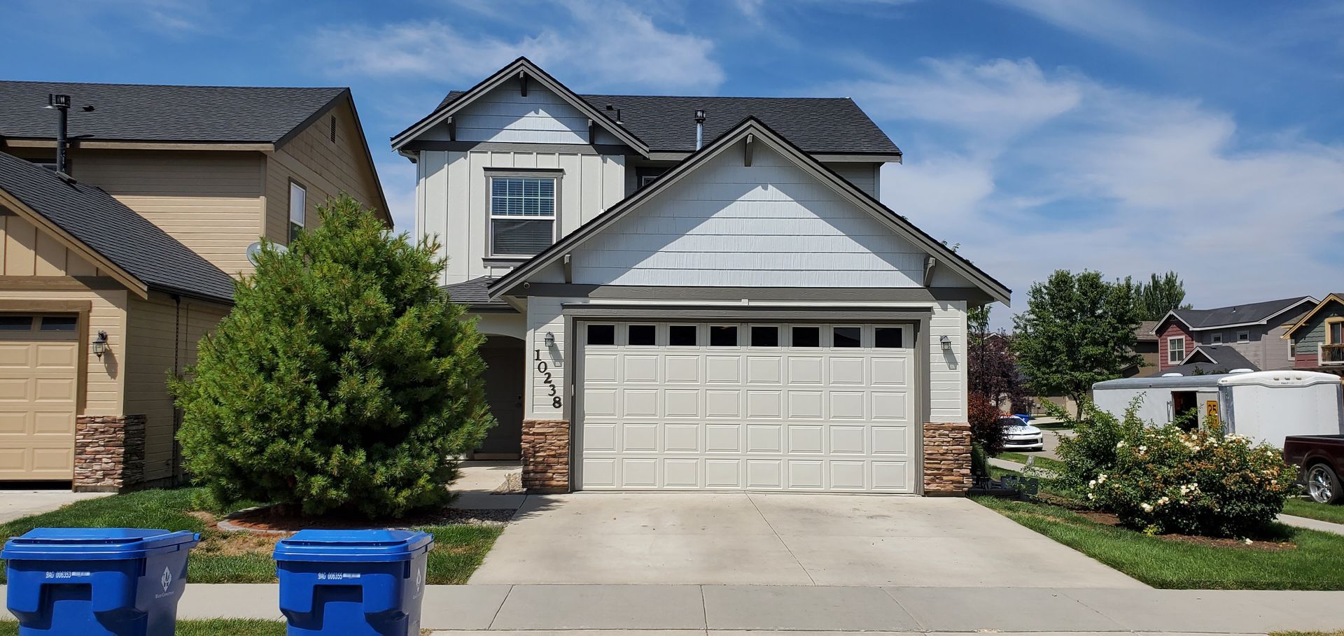 A two-story house with a light-colored exterior and a two-car garage, set against a blue sky.  Two blue trash cans are in front.