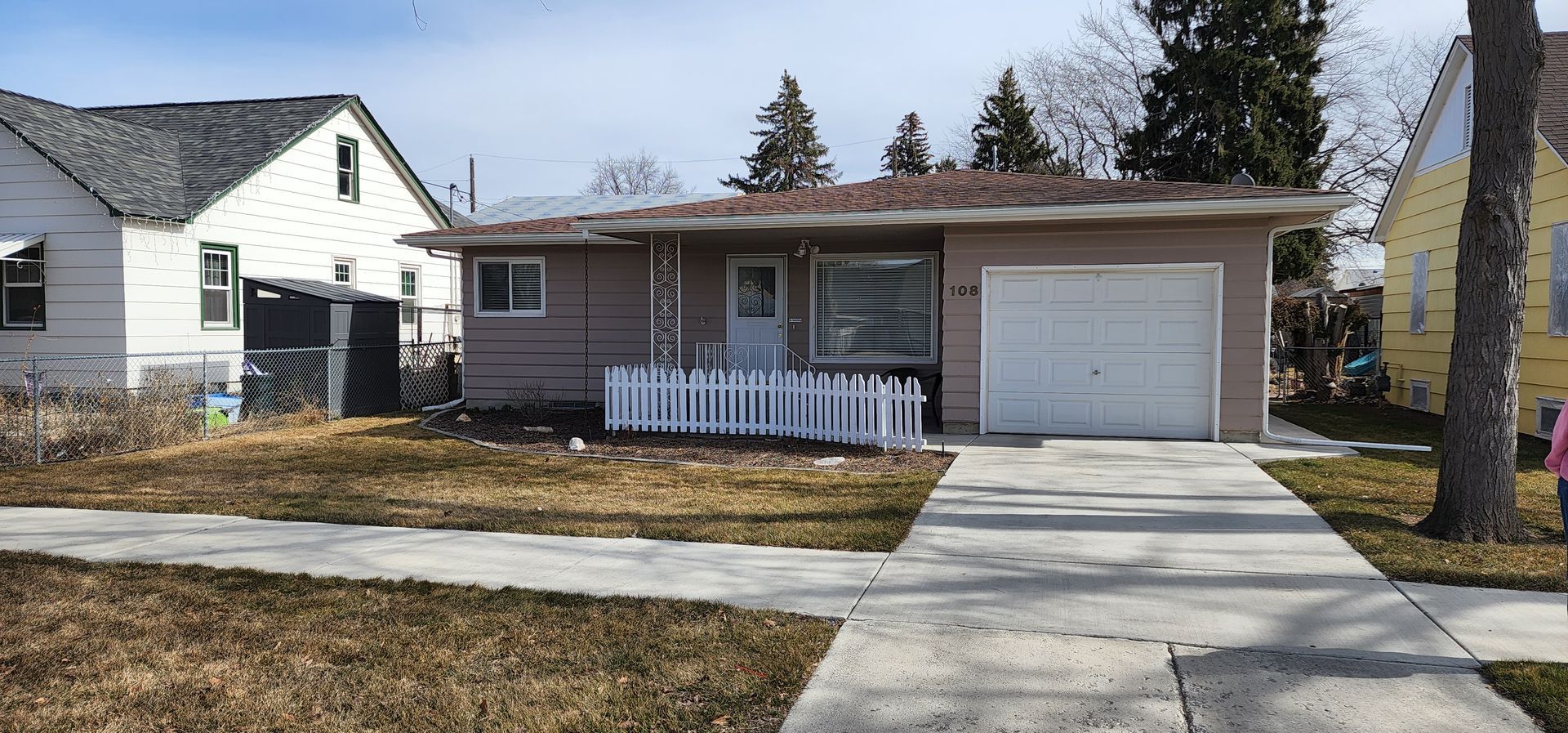 A single-story house with a picket fence in front, and a driveway leading to a garage. Other houses flank the central house.