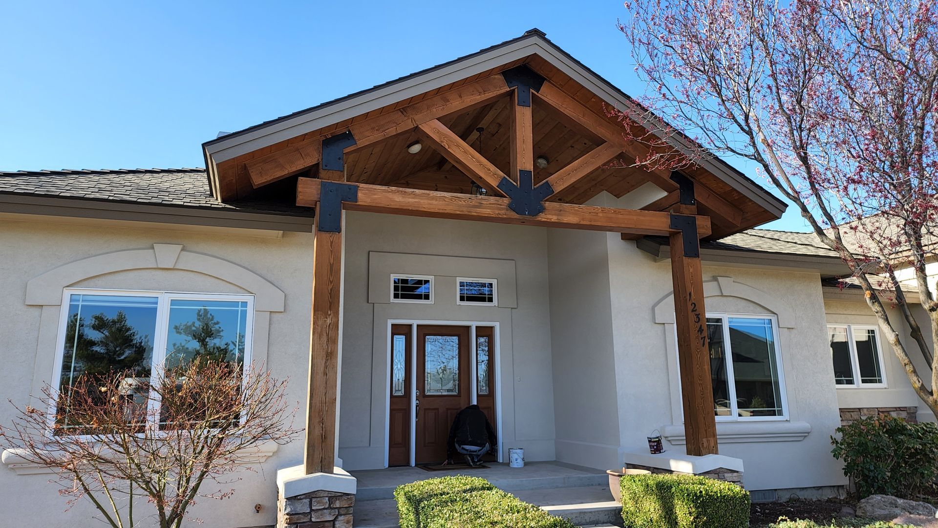 A house with a wooden-beam supported porch, stucco exterior, windows, and a brown door under a clear blue sky.