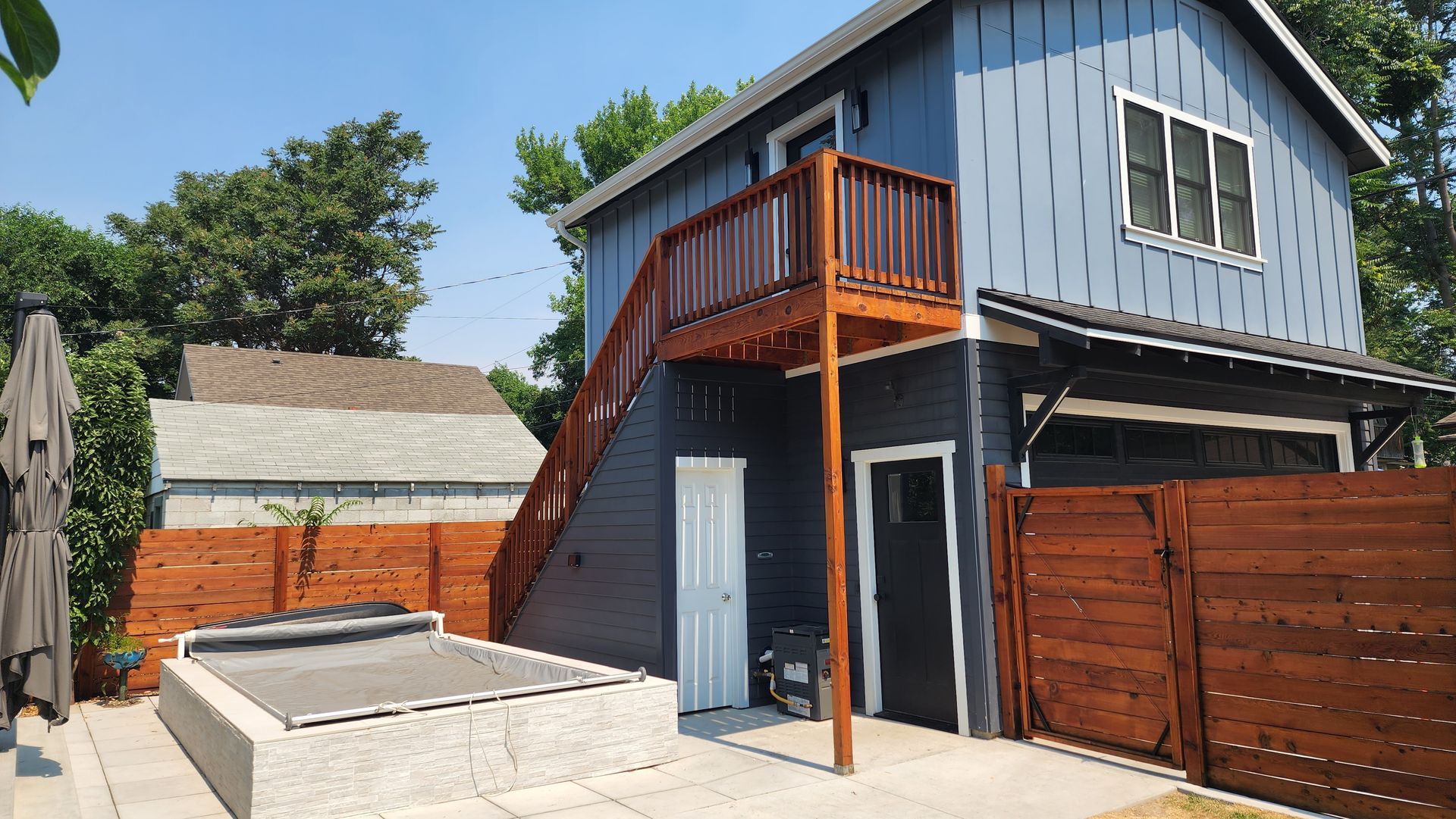 Two-story blue and brown building with a deck, stairs, and a garage. A wooden fence surrounds a concrete patio.