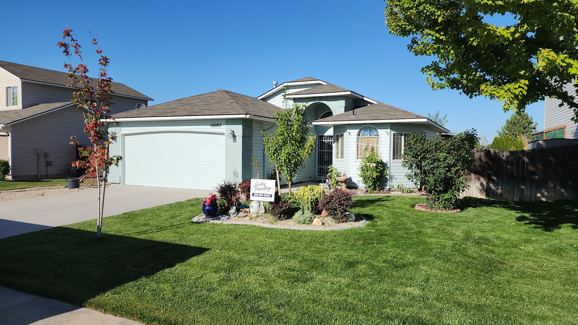 A light blue house with a gray roof and a well-manicured lawn on a sunny day. Landscaping includes small colorful flowers.
