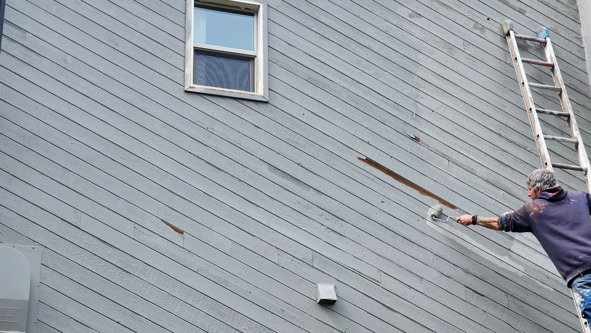Person on a ladder scraping grey siding on a house with a tool. A window is visible above.