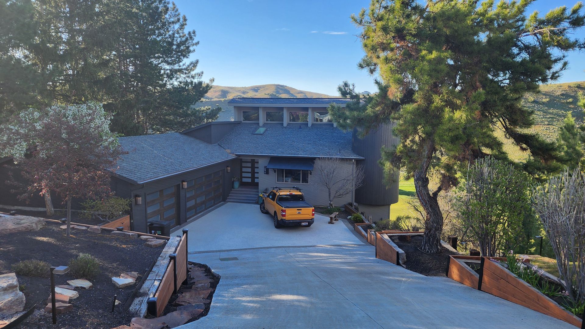 An orange truck parked in the driveway of a gray house with a dark roof, set against a hilly, green landscape.