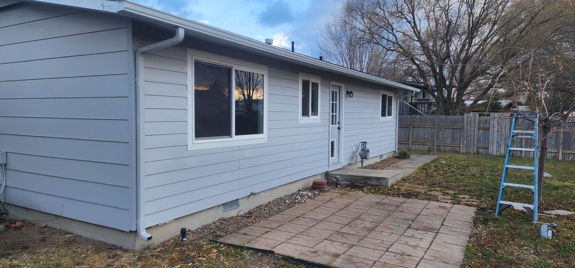 Exterior view of a light blue house with white trim and a yard with a wooden patio and a ladder.