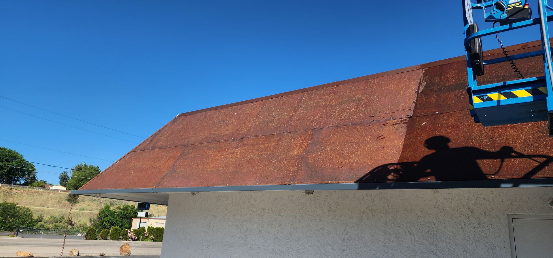A building's weathered roof is being worked on by someone in a blue lift on a clear, sunny day.
