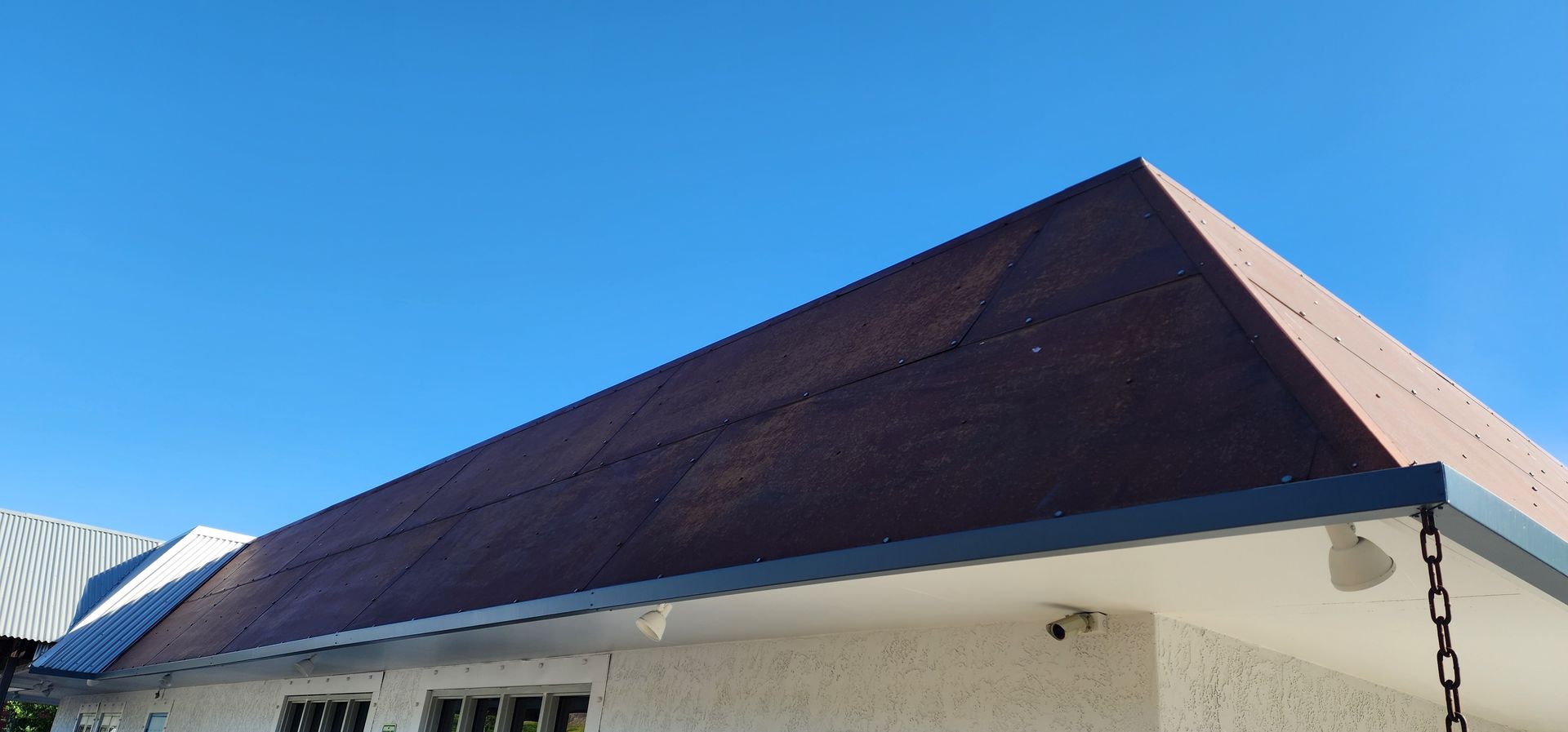 A building with a brown roof and a white exterior against a clear blue sky. A security camera is visible on the building.
