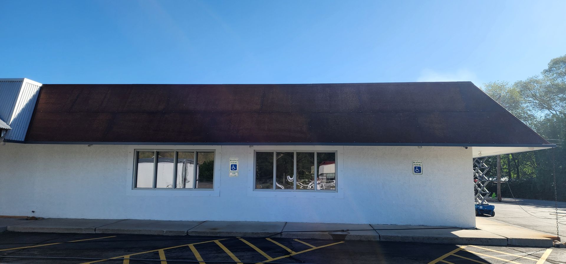 A white building with a brown roof. Windows and a parking lot are visible. The sky is blue.