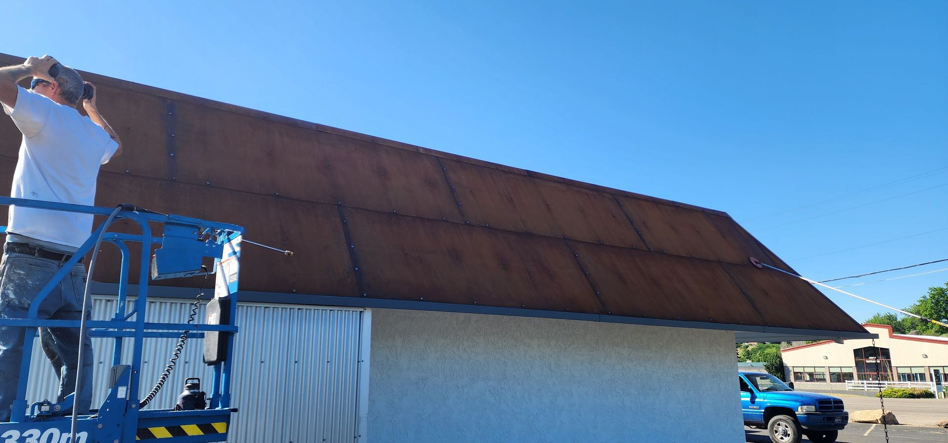 A person on a lift is working on a rusty brown metal roof. The setting is a sunny day with a blue sky.