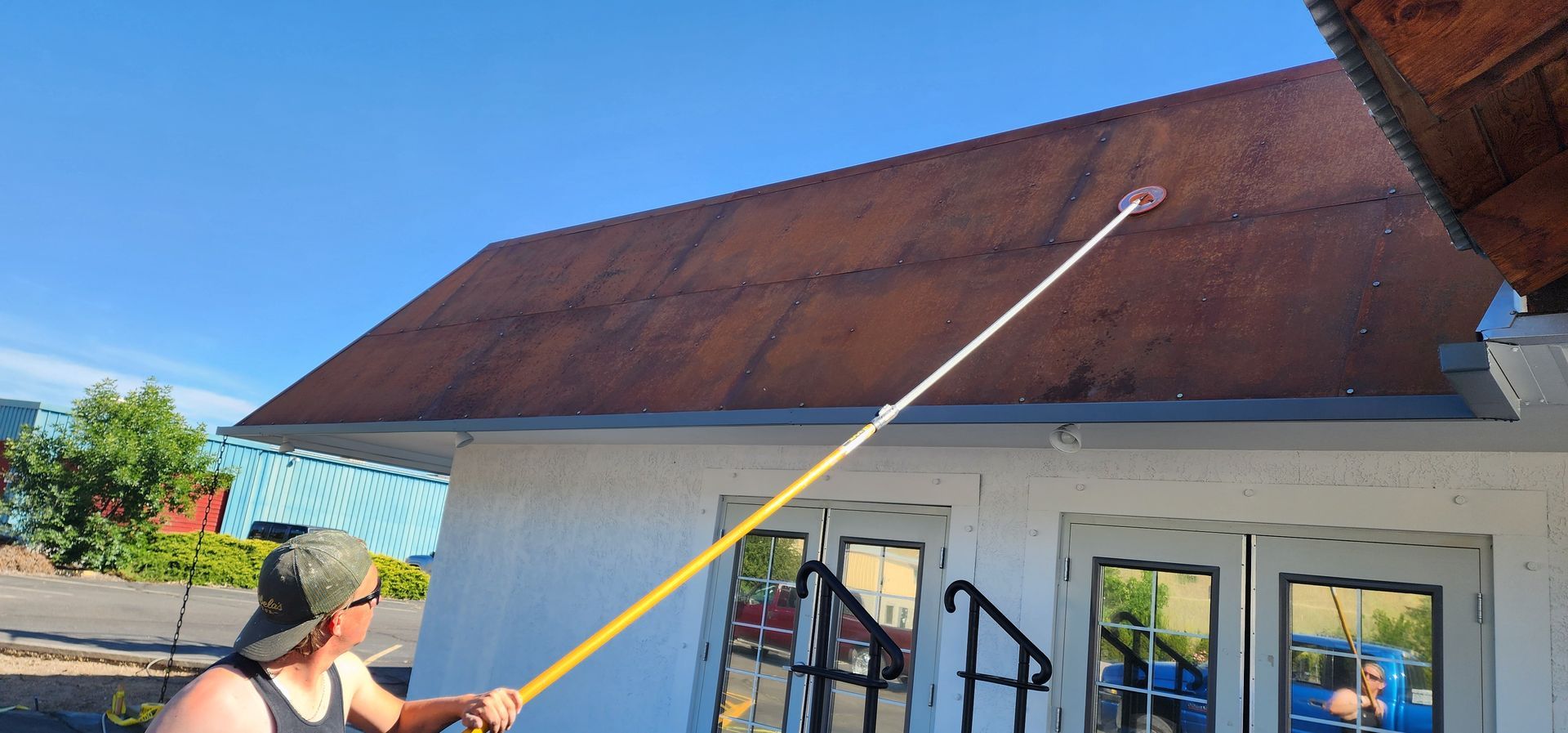 A person uses a pole to clean a rusty roof on a sunny day. The building is white with double doors.