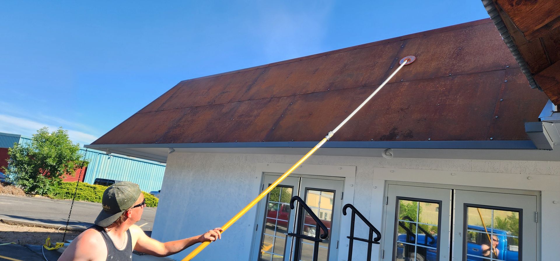 A person spraying a red roof with a long pole. The building is white with black handrails, and the sky is blue.