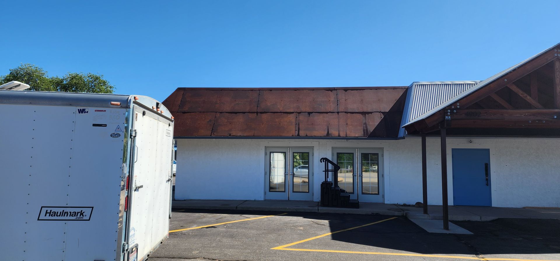 A white commercial building with a rusted brown roof and blue door. A delivery truck is parked in front of the building on a sunny day.