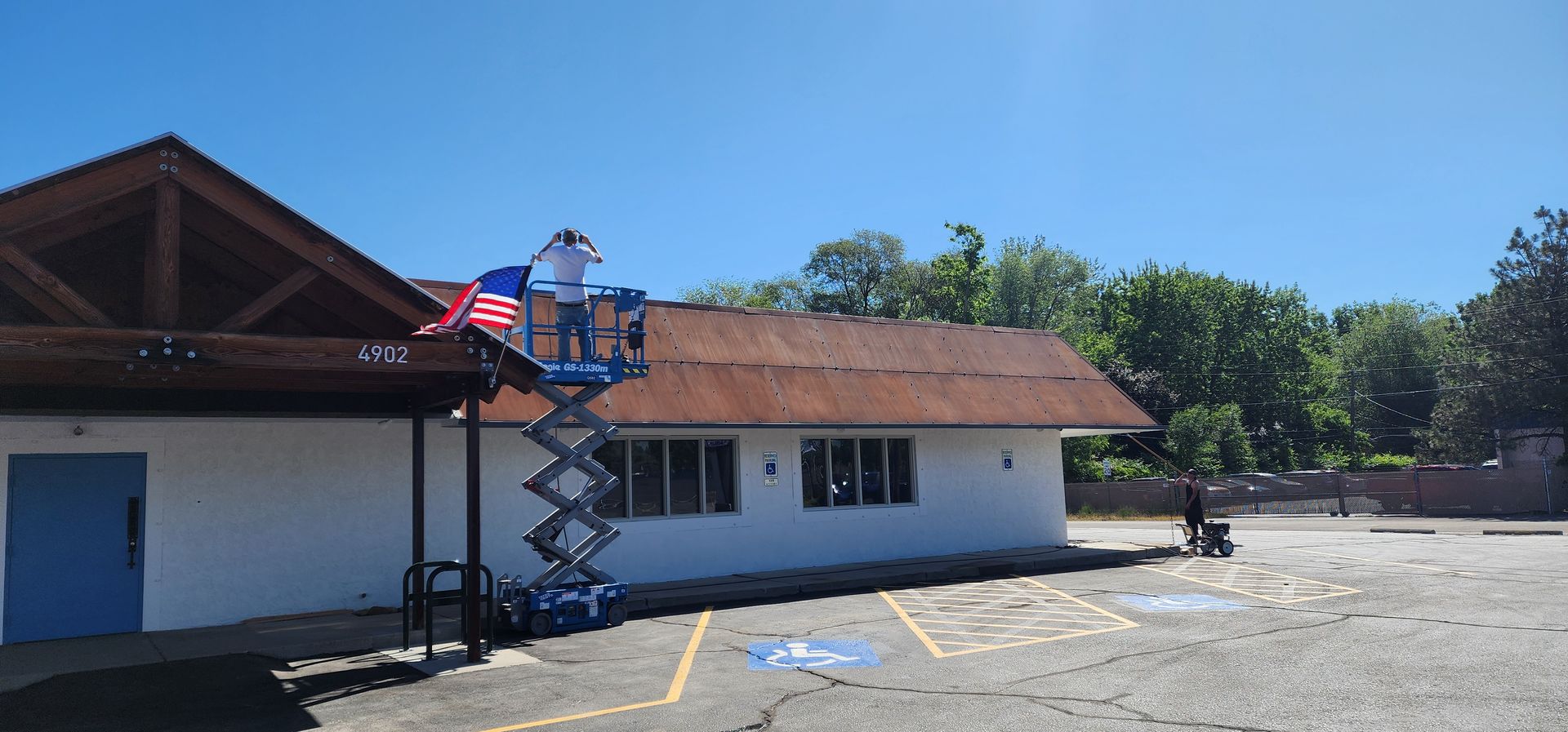 A person on a lift hangs an American flag on a building with a brown roof and blue door on a sunny day.