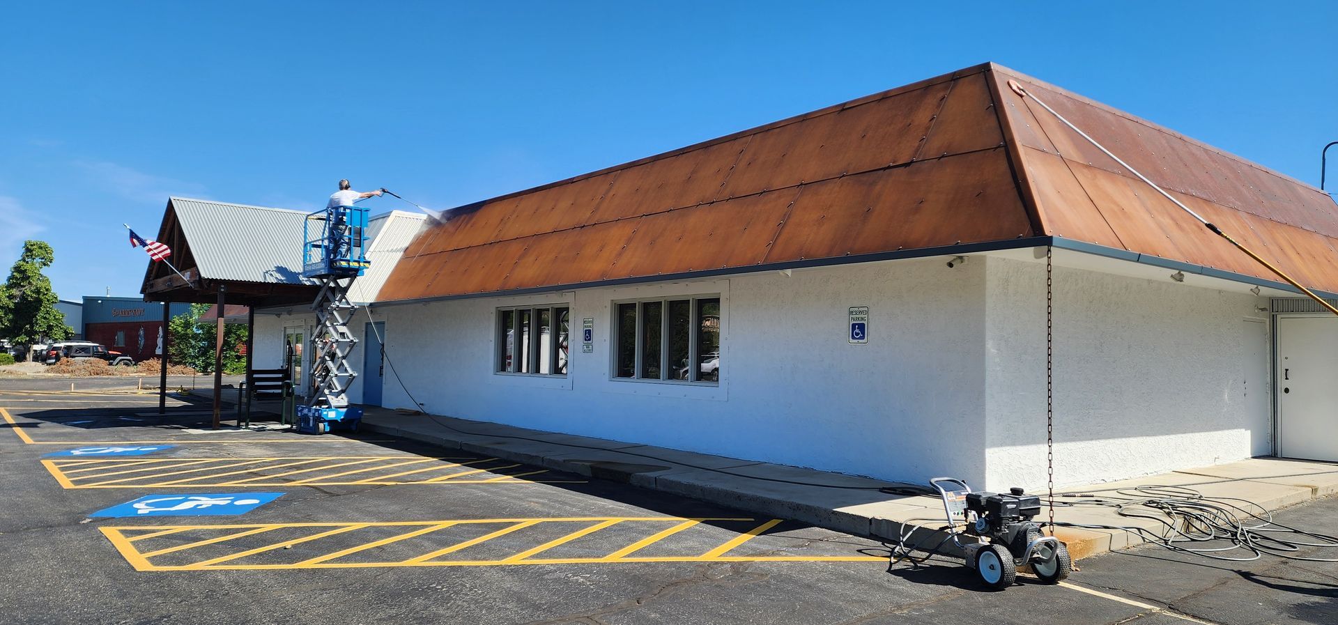 A person on a lift painting the white exterior of a commercial building with a rusted metal roof under a clear blue sky. Parking lot in the foreground.
