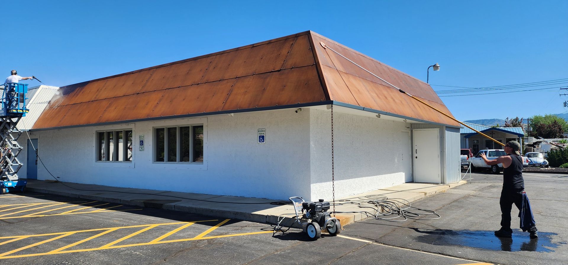 A person pressure-washing a white building with a rusty brown roof on a sunny day.  A lift is positioned near the building.