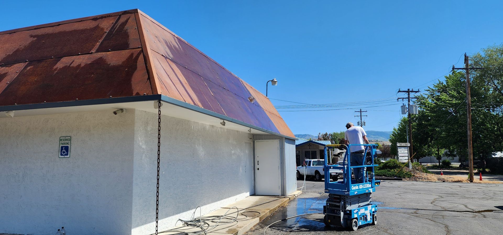 A person on a lift is washing a rusty-colored roof on a white building against a bright blue sky.