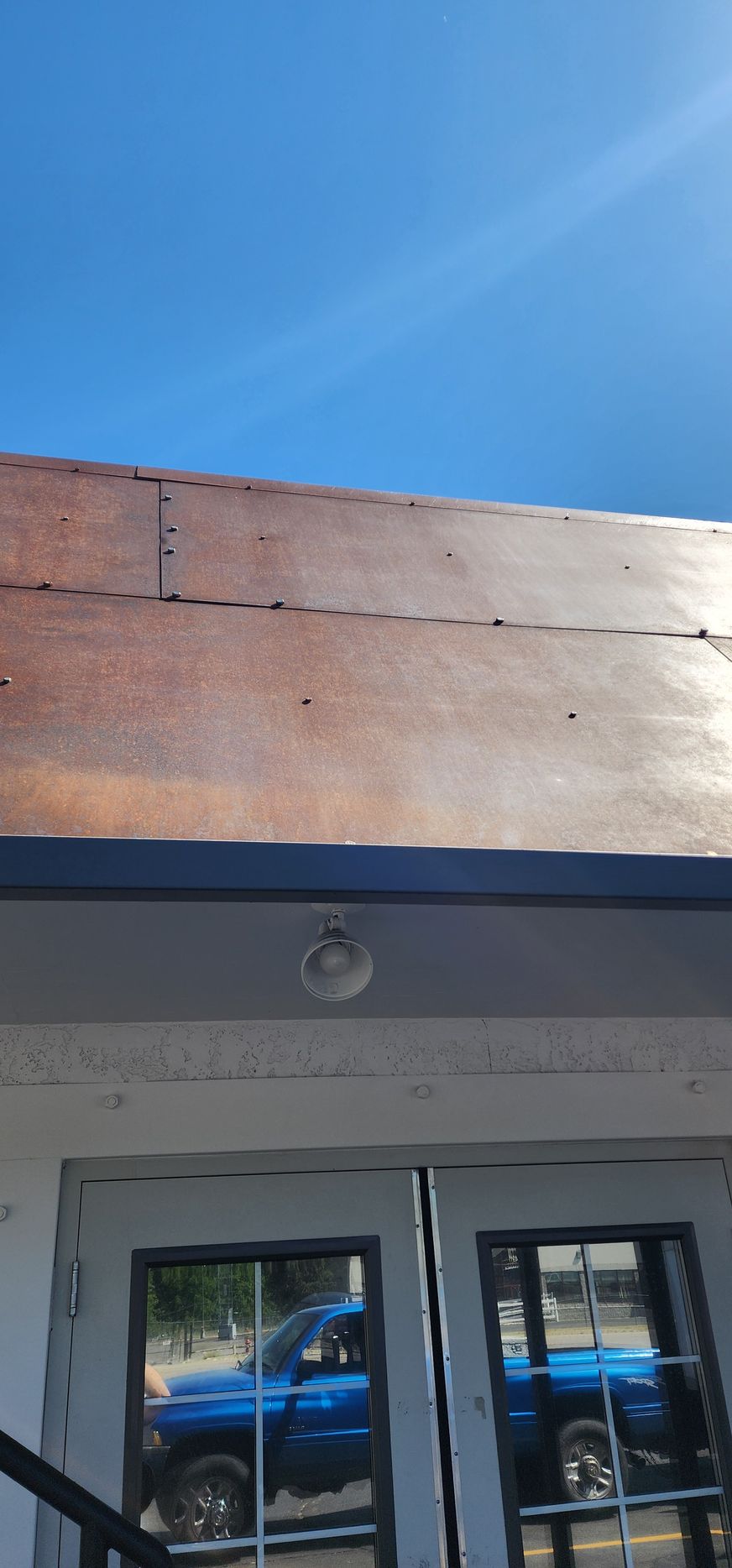 A building's weathered metal roof and white facade, with a blue truck reflected in the windows. The sky is bright blue.