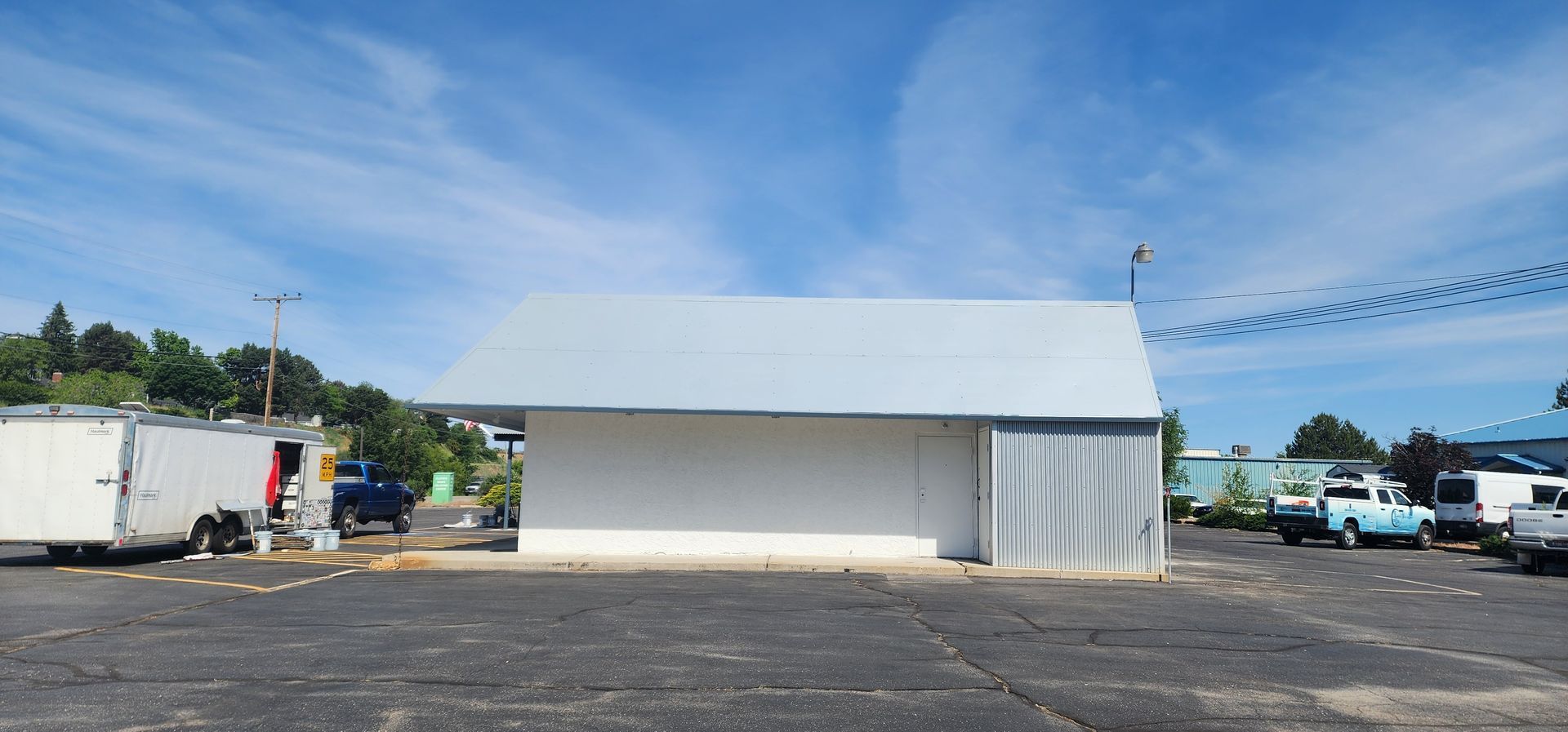 A white building with a gray roof and a parking lot. Trailers and vehicles are parked nearby, with a blue sky overhead.