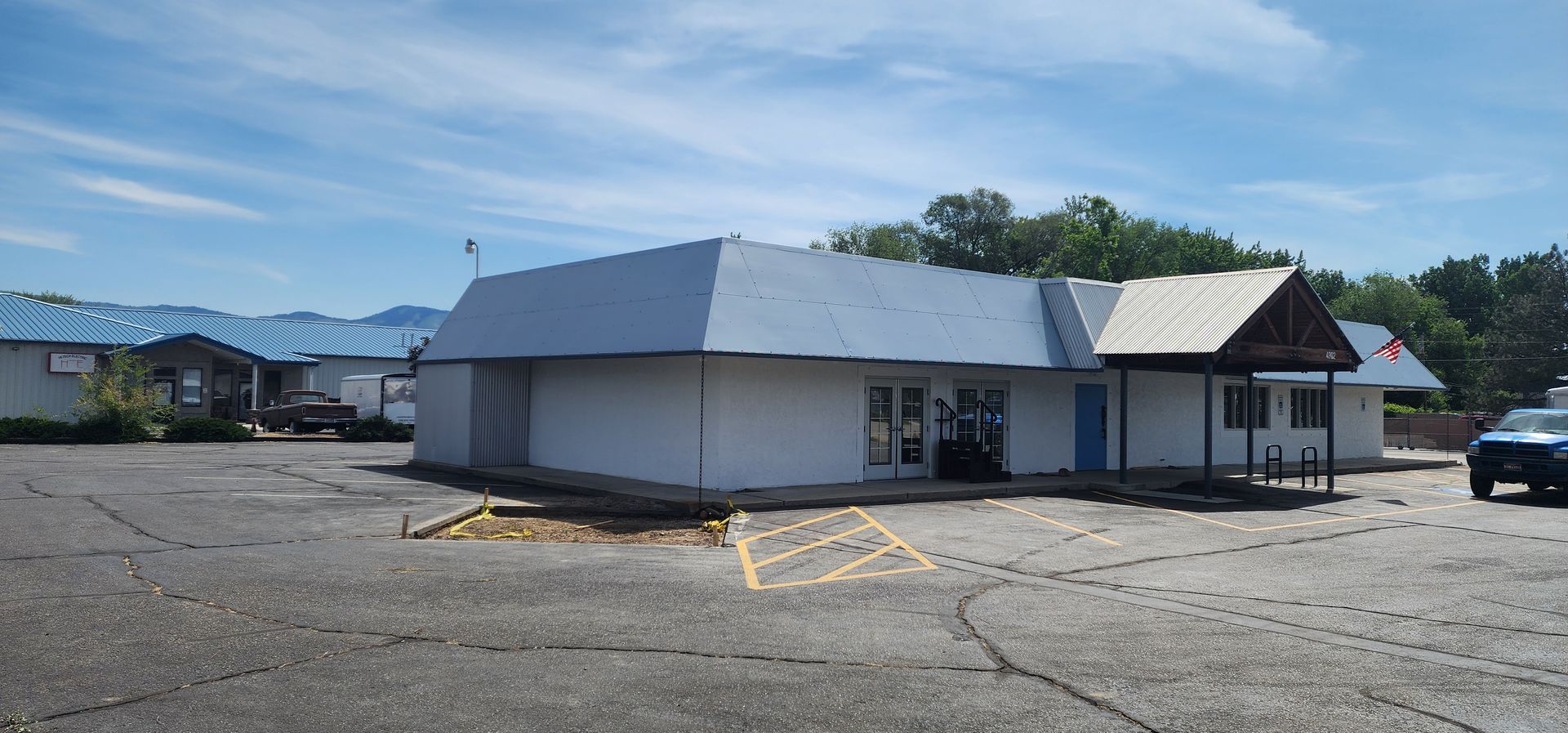 An abandoned building with a blue roof and white walls sits in a cracked parking lot on a sunny day.