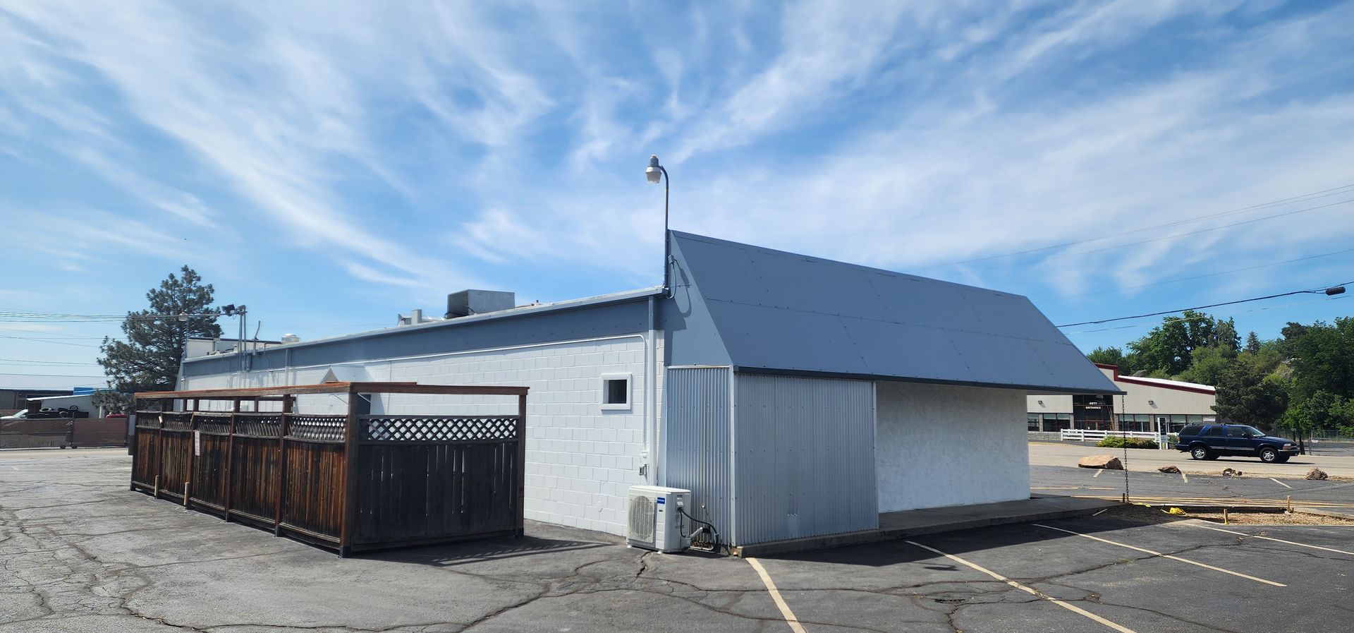 A white building with a blue roof under a partly cloudy sky. A brown wooden fence is in front of part of the building, and a black pickup truck is parked nearby.
