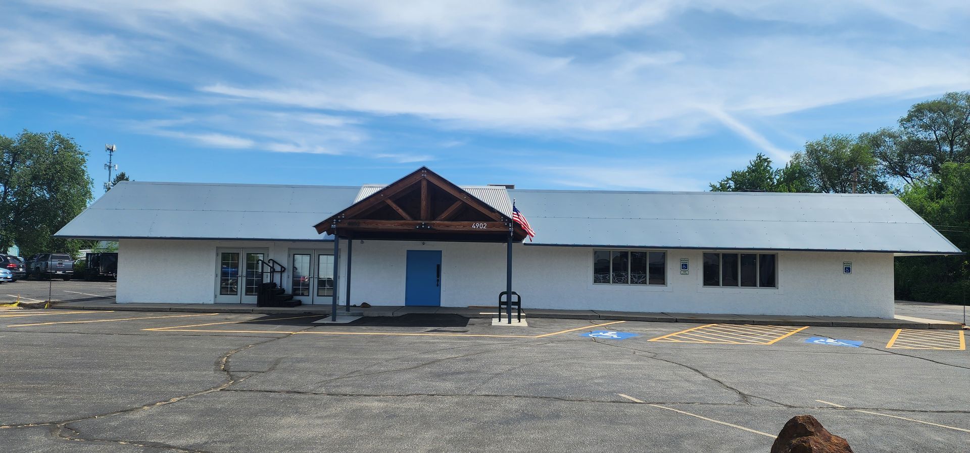 A white one-story building with a gray roof and blue door. A wooden awning covers the entrance. The sky is blue.
