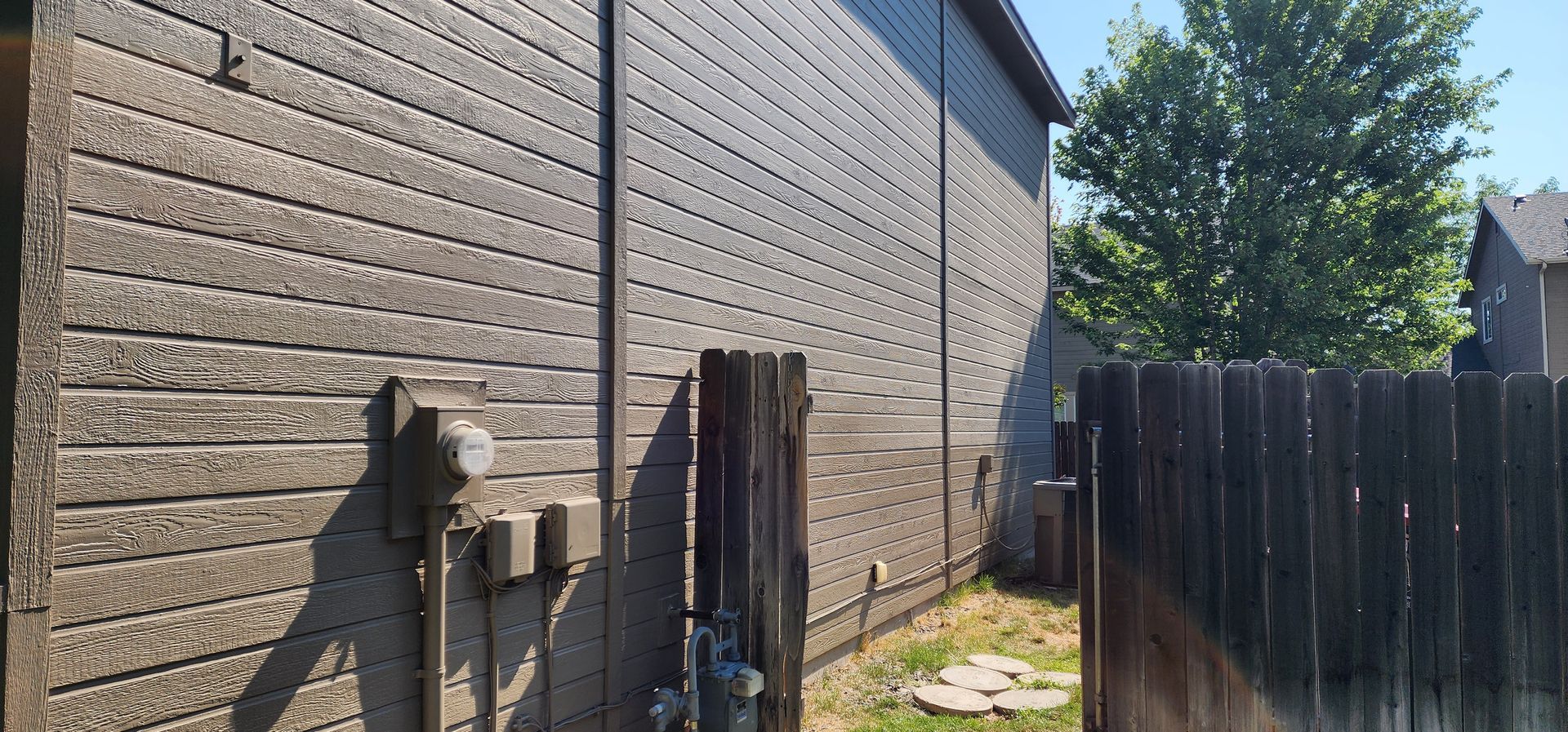 Exterior of a building with textured siding, a wooden fence, and a tree, bathed in sunlight.