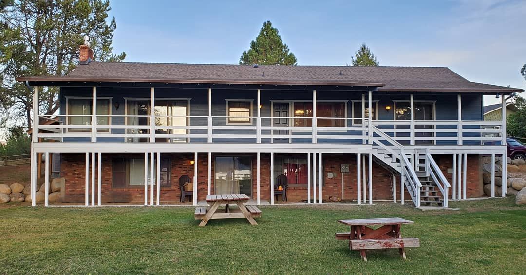 Two-story blue house with white railings and stairs, built on brick pillars. Picnic tables are in the green grass.