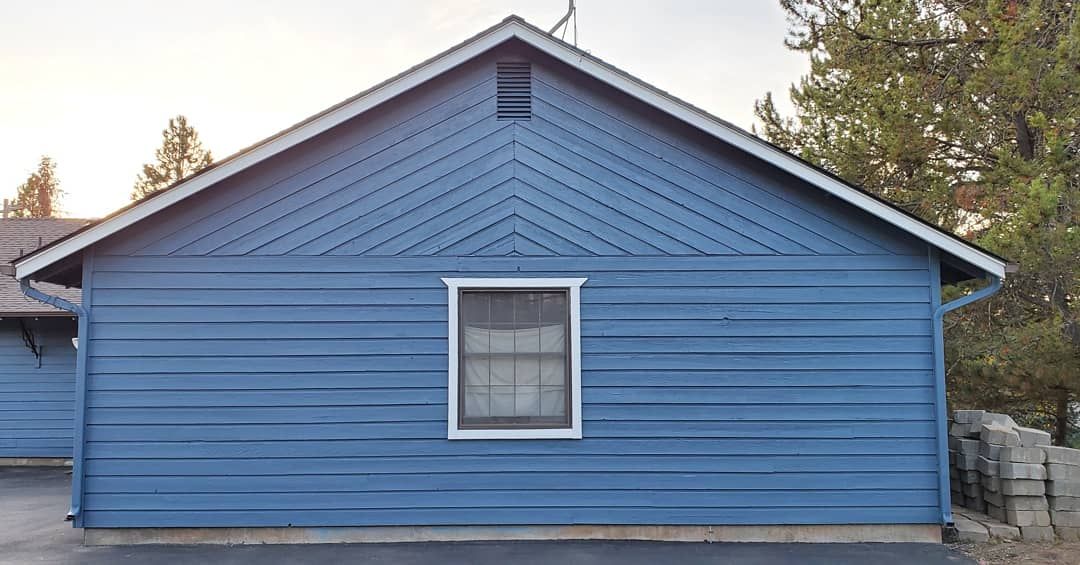 Blue-painted building exterior with white-framed window. Chevron detailing on the roof's peak; trees and a driveway are in the background.