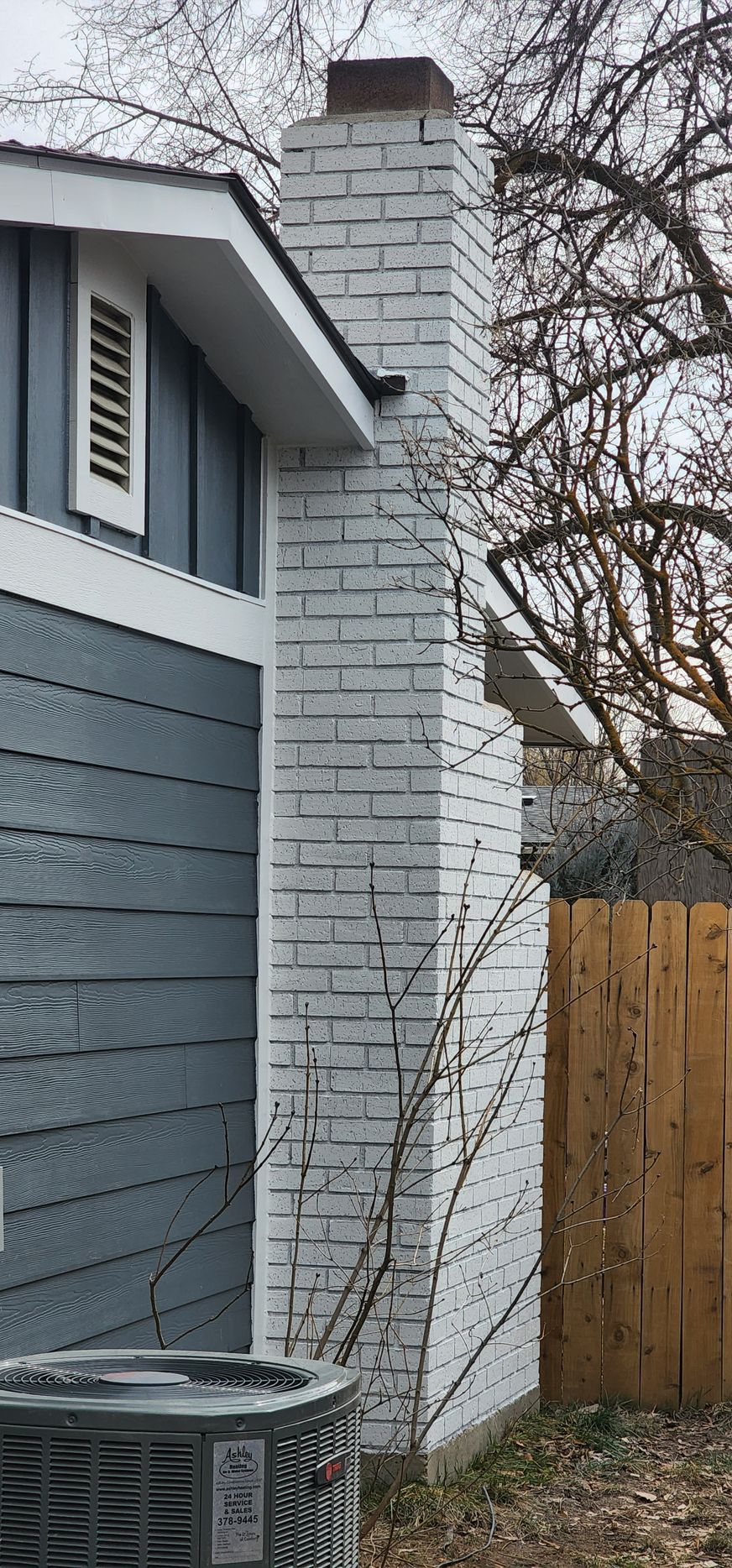 Exterior view of a house with a white brick chimney, gray siding, and a wooden fence in the background.