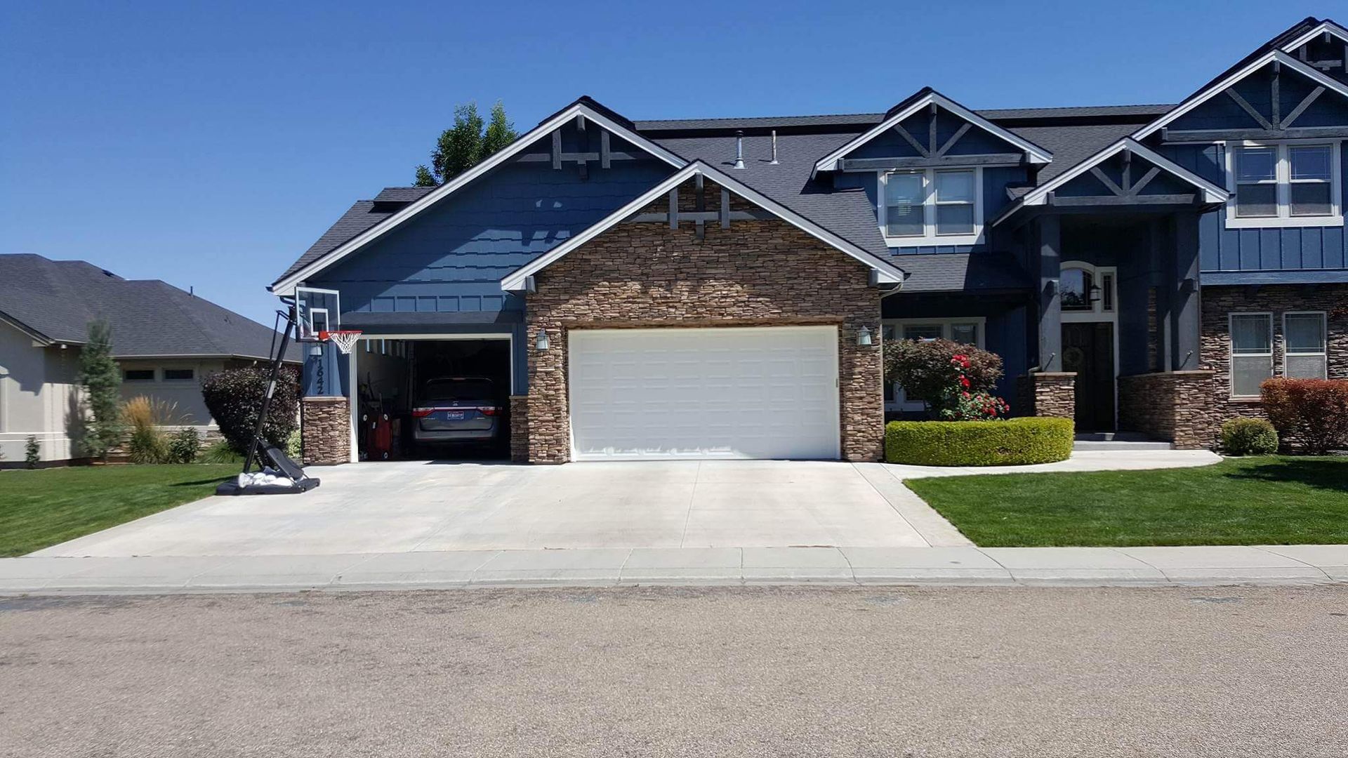 A two-story blue house with a stone garage door and a basketball hoop in the driveway on a sunny day.
