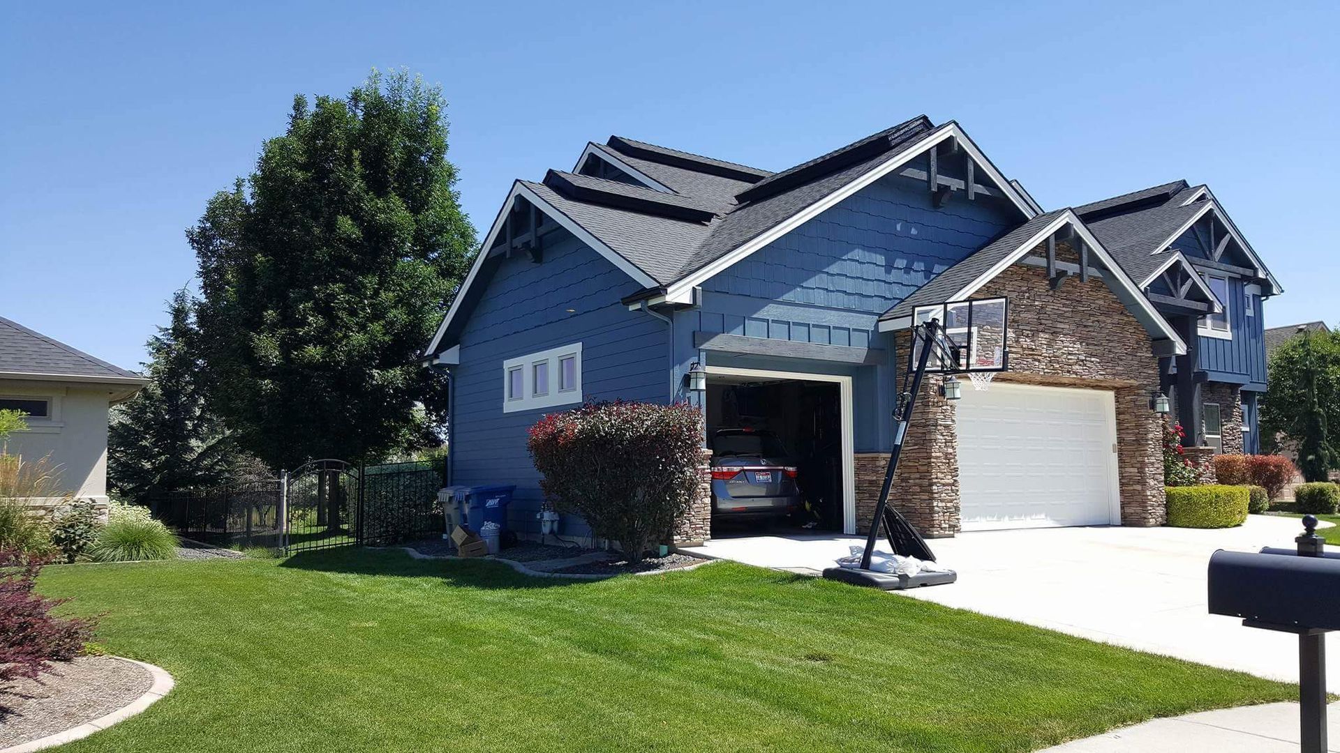 A blue two-story house with a stone-covered section and open garage door on a sunny day. A basketball hoop is in front.